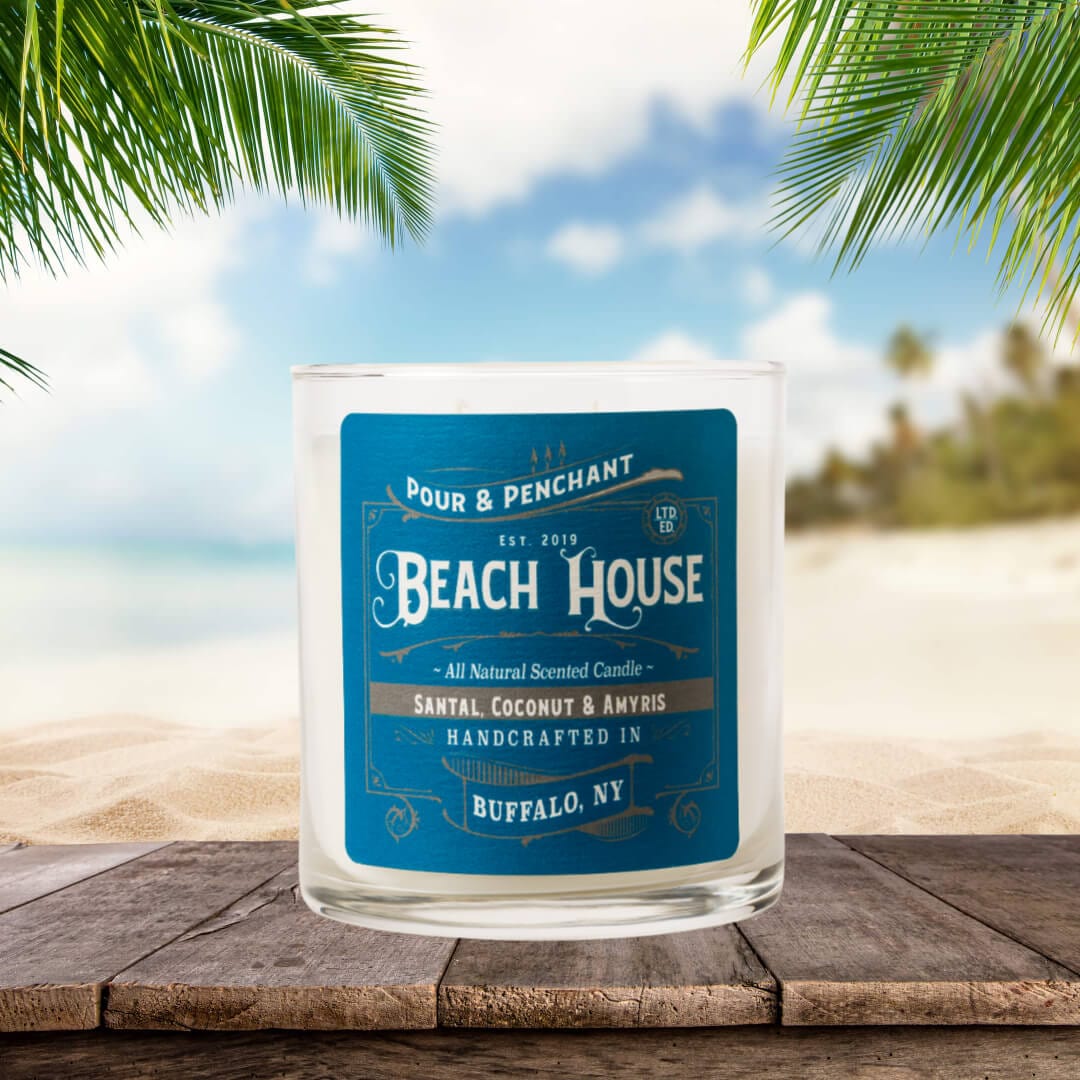 Pour & Penchant 10oz Beach House Scented Candle on a wooden boardwalk next to the ocean & palm trees